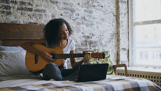 卷曲的非洲裔美国少女集中学习弹吉他使用笔记本电脑坐在家里的床上视频素材