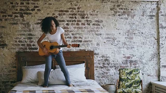 混合种族年轻有趣的女孩弹吉他和有乐趣跳舞在床上在家视频素材