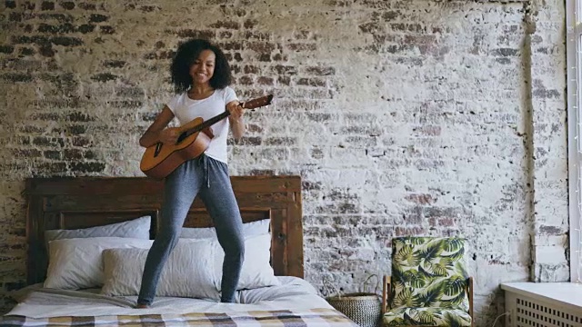 混合种族年轻有趣的女孩唱歌和弹吉他，而有乐趣跳舞在床上在家视频素材