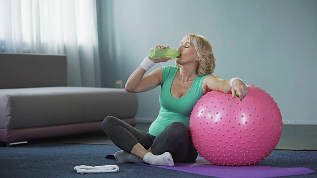 满意的老年女性锻炼后放松，在家饮水，运动视频素材