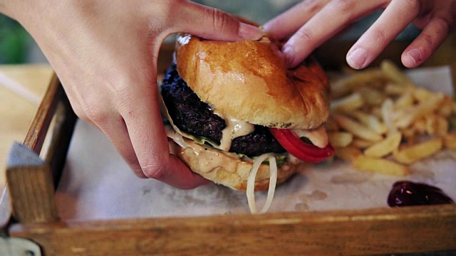 女人的手从一个木托盘上拿起一个大的美味汉堡，准备吃它。Slowmotion拍摄视频素材