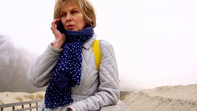 这位50岁迷人的老年女子在冬天的海滩上通过手机聊天的视频肖像视频素材