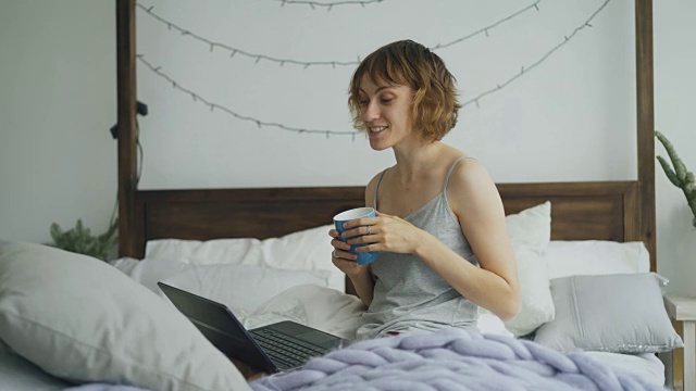 一个愉快的年轻女子坐在家里的床上用笔记本电脑摄像头和朋友视频聊天视频素材