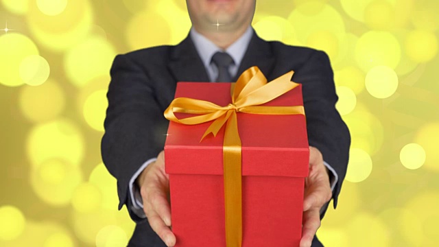 一个穿西装打领带的男人拿着一个红色礼品盒。男人给予礼物。一个手里拿着礼物的商人。背景抽象金色闪光闪烁，圈光散焦。视频素材