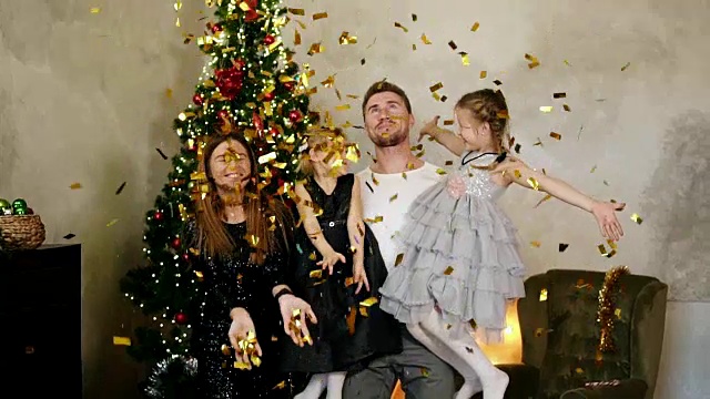一个快乐的四口之家在圣诞树旁抛起金色的五彩纸屑。可爱的妈妈，爸爸和两个女儿一起庆祝圣诞节视频下载