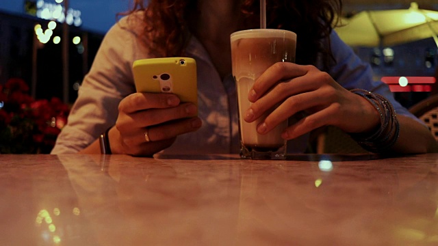 一名年轻女子喝了一杯鸡尾酒，在一个夏日的咖啡馆晚上使用手机视频下载