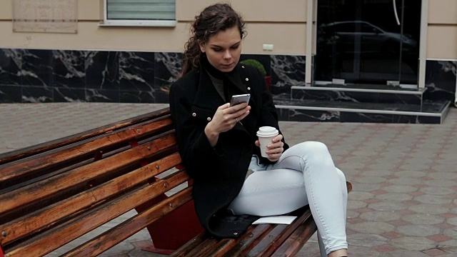 穿着外套的年轻女子坐在长凳上，喝着咖啡，用着手机。缓慢的运动。视频下载