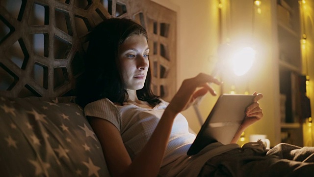 年轻微笑的女子晚上躺在家里的床上用平板电脑网购视频素材