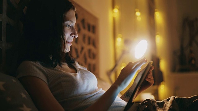 快乐微笑的女人晚上躺在家里的床上用平板电脑分享社交媒体视频素材