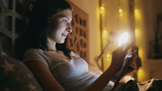 年轻微笑的女孩晚上躺在家里的床上使用平板电脑视频素材