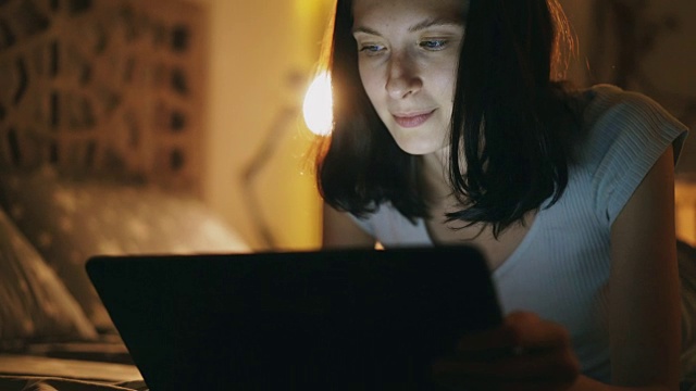 年轻迷人的女人在晚上躺在家里的床上使用平板电脑的特写视频素材