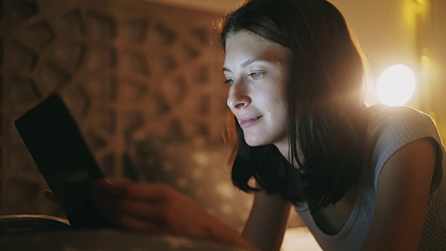 年轻漂亮的女人在晚上躺在家里的床上使用平板电脑的特写视频素材