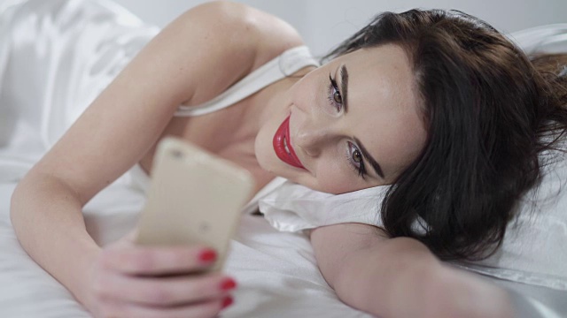 白色床单上躺着一个穿着内衣手拿智能手机的漂亮女人视频素材