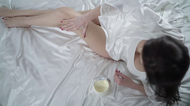 美丽的女人坐在床上，在光滑的腿上涂霜。腿皮肤护理。视频素材