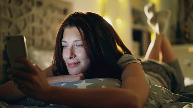 年轻微笑的女子晚上躺在床上用智能手机分享社交媒体视频素材