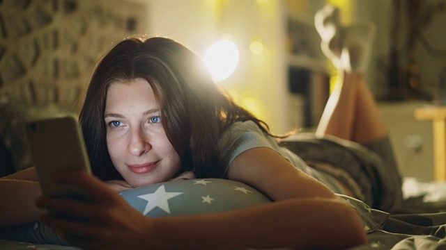 迷人微笑的女人晚上躺在床上用智能手机分享社交媒体视频素材