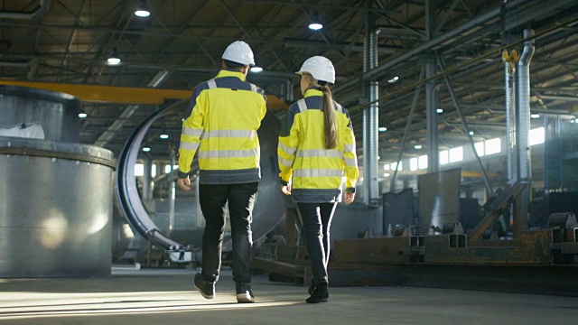 男工程师和女工程师在重工业制造厂边走边讨论的背影照片。大型金属制品结构，管道元素躺在周围。视频素材