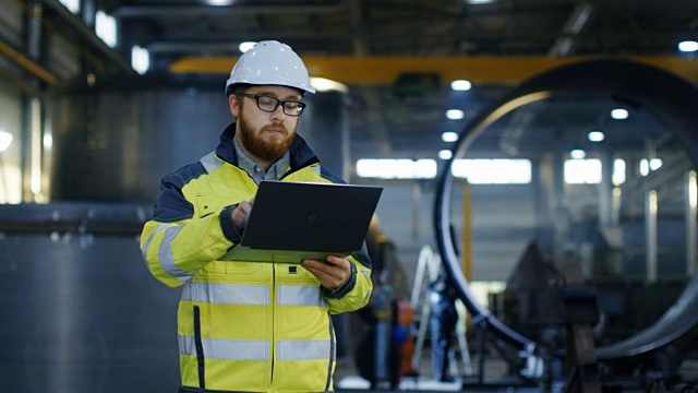 工业工程师戴安全帽穿安全外套使用笔记本电脑。他在重工业制造工厂工作，各种金属加工工艺正在进行中。视频素材