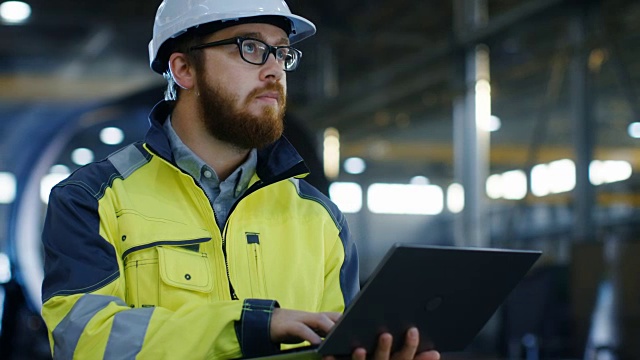 工业工程师戴安全帽穿安全外套使用笔记本电脑。他在重工业制造工厂工作，各种金属加工工艺正在进行中。视频素材