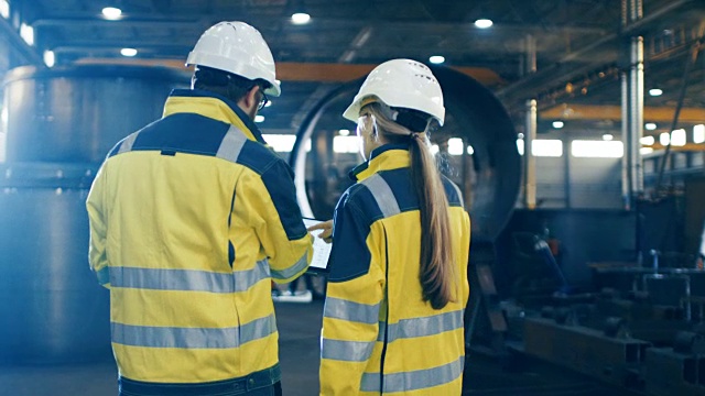 男性和女性工业工程师使用笔记本电脑和讨论在重工业制造工厂。他们戴安全帽和安全外套。背景:大型金属制品结构，管道部件。视频素材