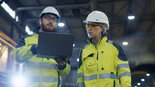 男性和女性工业工程师使用笔记本电脑和讨论在重工业制造工厂。他们戴安全帽和安全外套。视频下载