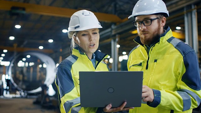 在重工业工厂里，女工业工程师拿着笔记本电脑和项目经理讨论。他们戴安全帽和安全外套。正在进行的焊接/金属加工。视频素材