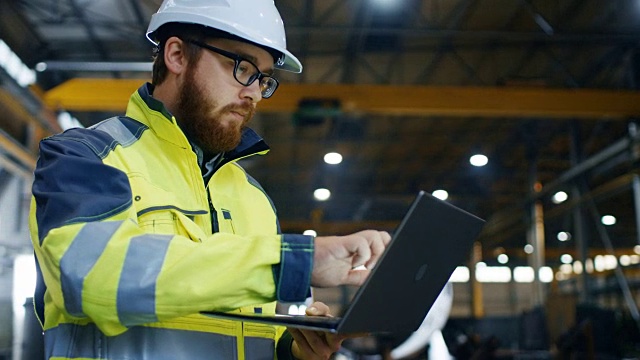 工业工程师戴安全帽穿安全服使用触屏笔记本电脑。他在重工业制造厂工作。焊接/金属加工工艺正在进行中。视频素材