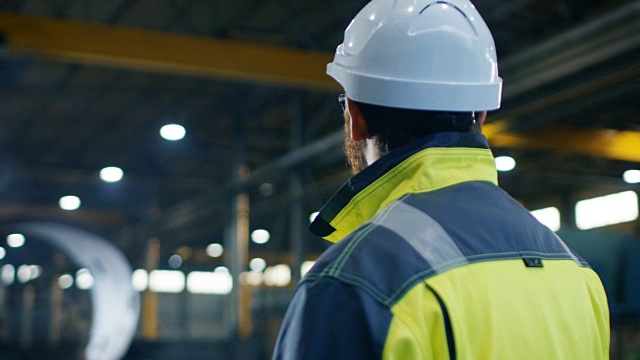 戴安全帽、穿安全服的工业工程师在大型重工业制造工厂四处查看。弧查看录像。视频素材