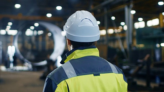 戴安全帽、穿安全服的工业工程师在大型重工业制造工厂四处查看。弧查看录像。视频素材