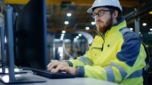 工业工程师在工作空间用个人电脑工作。他戴着安全帽和安全外套，在重工业制造工厂的主车间工作。视频素材