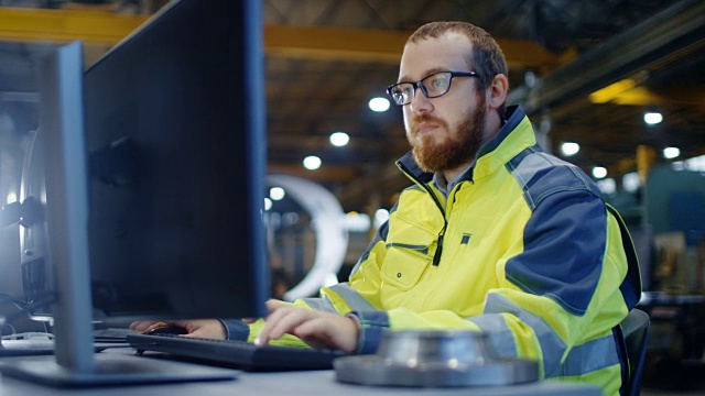 工业工程师在工作空间用个人电脑工作。他穿着安全夹克，在重工业制造工厂的主车间工作。视频下载