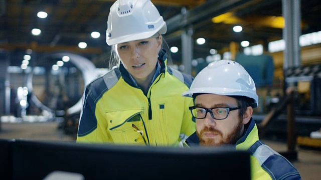 男性工业工程师工作在个人电脑上，而女性经理谈论项目细节。他们在重工业制造工厂工作。视频素材