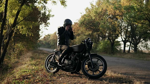 一个穿着黑色皮夹克的年轻人坐在他的摩托车上，戴上黑色的头盔去森林旅行。慢动作镜头视频素材