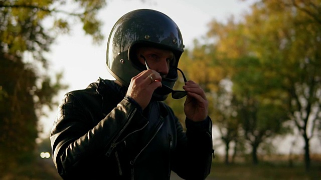 一个年轻人穿着黑色皮夹克和头盔戴着太阳镜，而坐在他的摩托车上准备开始他的旅程在森林。慢动作镜头视频下载