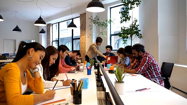 多样化的创意团队在办公室工作，每个人都在完成自己的任务，而领导正在和一个使用笔记本电脑的人交谈视频素材