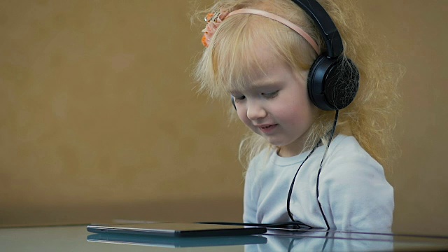 慢镜头:一个三岁的小女孩戴着大耳机在平板电脑上听音乐。视频素材