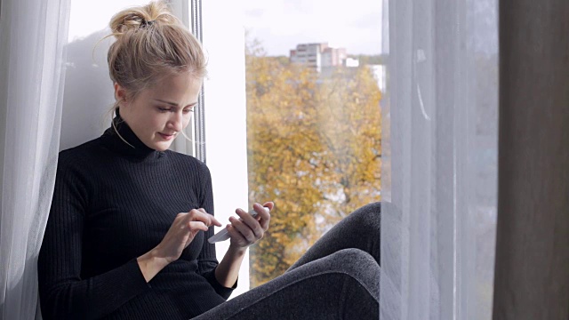 年轻女子坐在窗台上玩智能手机视频下载