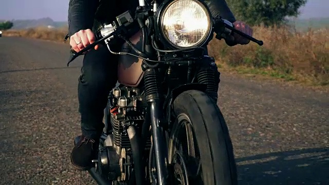 前面的观点，一个不认识的人在黑色皮夹克骑摩托车在一个乡村道路上持有一个把手。大灯打开了视频素材