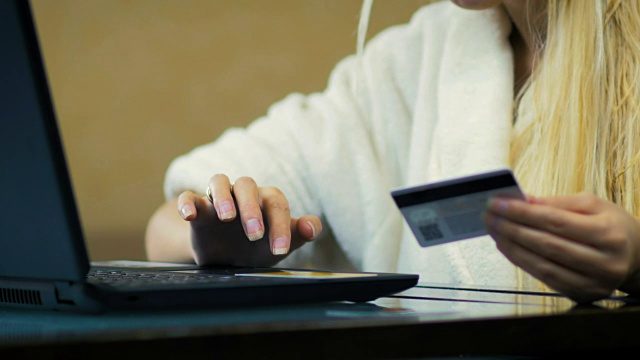 穿着白色长袍的金发女郎手里拿着一张信用卡，在家用笔记本电脑在网上商店购物。视频下载