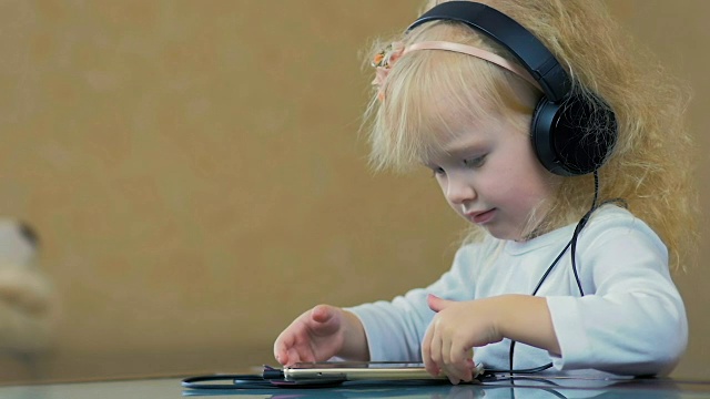 慢镜头:3岁的小女孩戴着耳机在家里用智能手机听音乐。视频素材