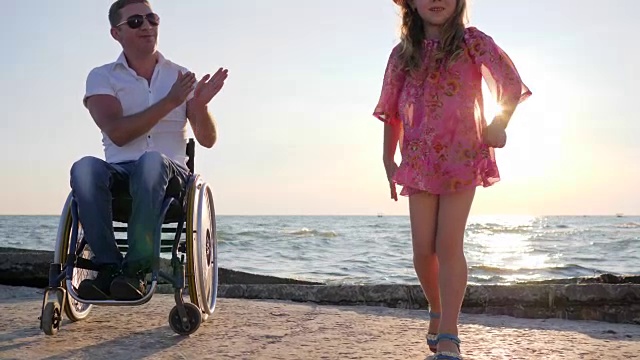 坐在轮椅上的残疾人在海边的背光慢镜头中看着跳跃的孩子为女儿鼓掌视频下载