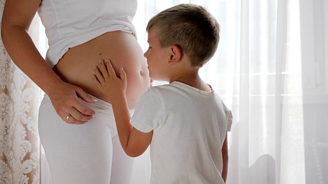 白色窗帘背景下，好奇的男孩饶有兴趣地倾听着孕妇的肚子视频下载
