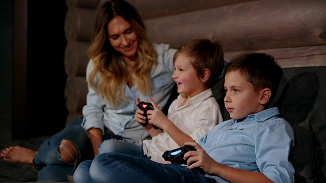 母亲和两个儿子坐在他家的沙发上用无线操纵杆玩电子游戏。快乐的人在房子里视频下载