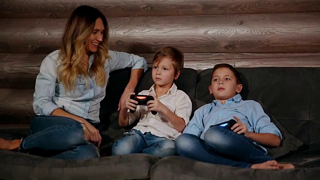 母亲和两个儿子坐在他家的沙发上用无线操纵杆玩电子游戏。快乐的人在房子里视频下载