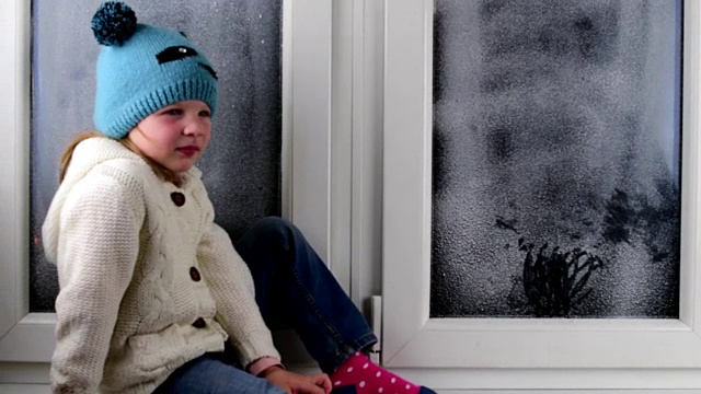 小女孩坐在结冰的窗户后面的窗台上。低的自由度。视频下载