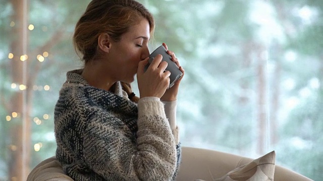 舒适的冬天的生活方式。年轻快乐的女人喝着咖啡，穿着针织毛衣，坐在家里的大窗户与冬天的雪背景视频下载