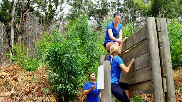 女教练协助健康女子在障碍赛4k期间爬过木墙视频素材