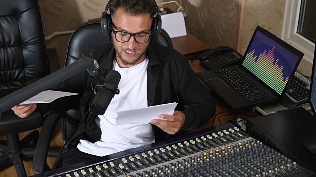 男电台主持人戴着眼镜，用一张纸对着电台演播室附近设备的麦克风讲话视频素材