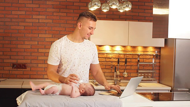一位年轻的父亲一边在厨房给小儿子喂奶，一边在用笔记本电脑工作视频下载