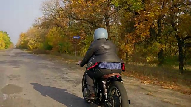 一个男人在灰色头盔和皮夹克和格子衬衫骑摩托车在柏油路在阳光明媚的一天秋天。路的周围长着棕色和黄色叶子的树视频素材
